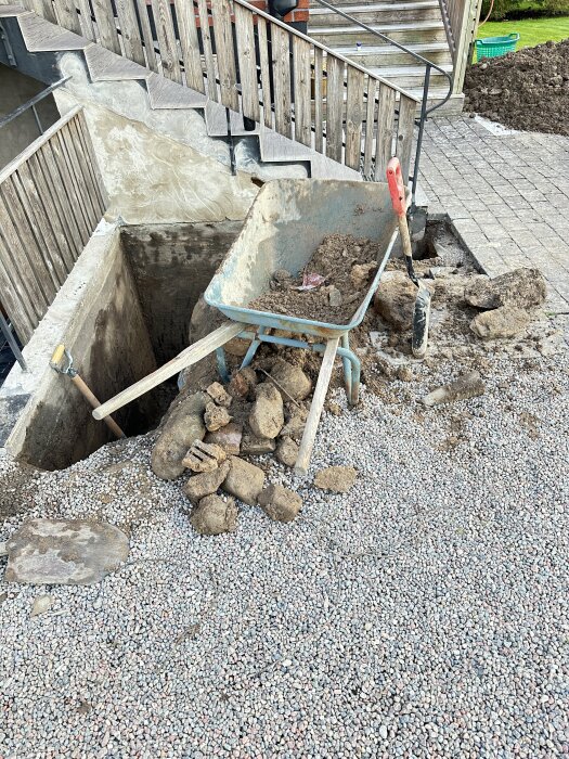 Uppgrävd plats vid en husvägg med en skottkärra fylld med jord och stenar; omgivande yta täckt av grus och sten, trappa synlig i bakgrunden.