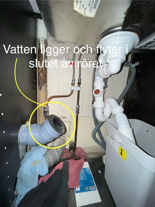 Avloppsrör under diskbänk med gul markering visar ett igensatt rör där vatten ansamlas. Rensband ligger bredvid samt tyg och plastbehållare närvarande.