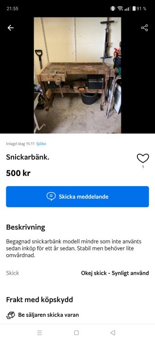 Begagnad snickarbänk i trä med hyllplan under, fylld med verktyg och redskap; spade står lutad mot väggen och en svart plasthink syns under bänken.