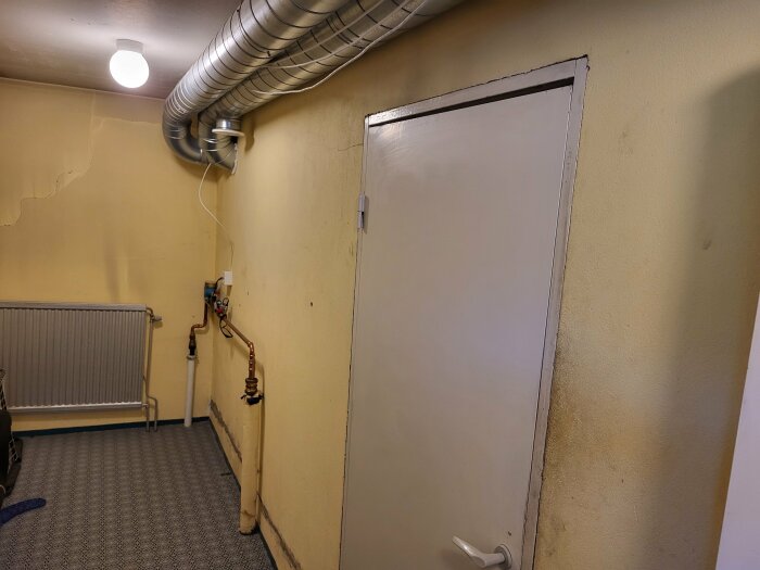 Tvättstuga med en dörr till pannrum, synliga rörinstallationer och radiator på en gul vägg. En spricka syns i väggen. Taklampa och ventilationsrör i taket.