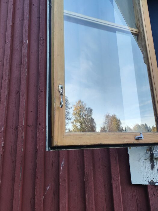 Öppet fönster med träkarm på en röd träfasad. Utsikten inkluderar träd och en klar himmel. En gummilist är installerad på karmen för tätning.