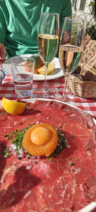 Lunch på en uteservering med råbiff, två glas champagne, vatten, lax, rostat bröd och citron på en rutig duk i sommarväder.