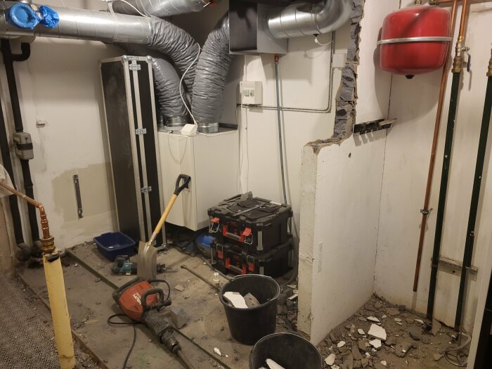 Ett pannrum under renovering med vattenrör, ventilationskanaler, en rödmålad expansionstank och påbörjad rivning av en innervägg. Verktyg och maskiner ligger på golvet.