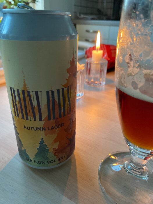 En burk med "Rainy Days Autumn Lager" och ett glas med en mörk öl på ett bord med ett tänt ljus i bakgrunden.