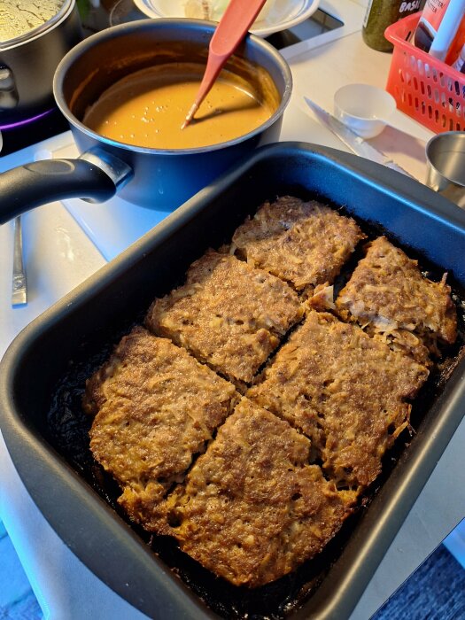 En bakform med köttfärslåda skuren i bitar och en kastrull med brun sås på ett köksbord.