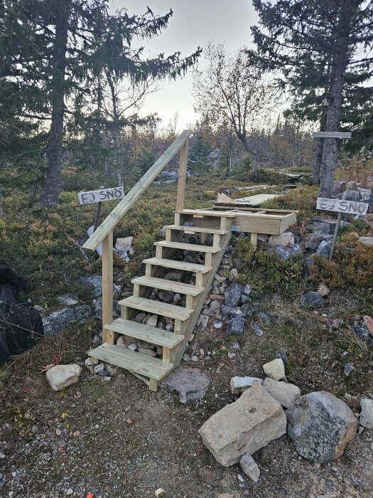 Trätrappa utomhus byggd på en stenig kulle, omgiven av granar. Handgjorda skyltar som säger "Ej snö" sitter på trädet och trästommen.