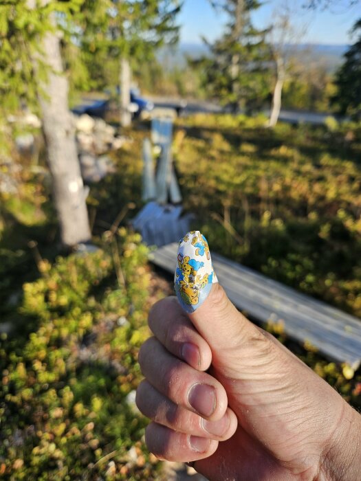 Tummen på en hand med ett barnplåster. Oskarp trädgårdsbakgrund med träbitar och byggmaterial synliga.