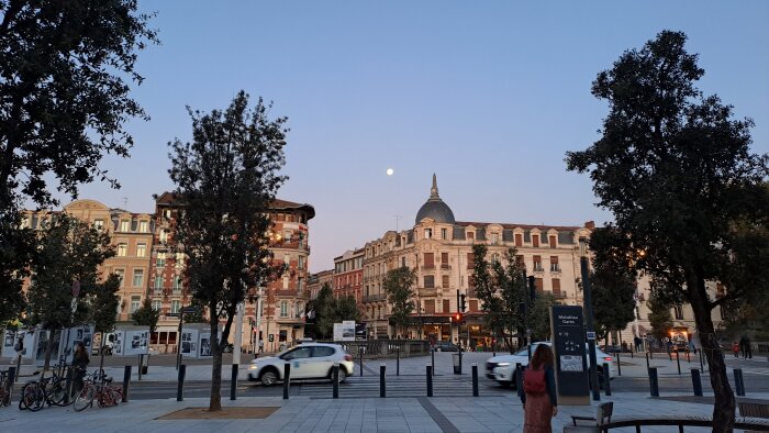 Stadsvy i Toulouse tidigt på morgonen med äldre byggnader, träd, cyklar och en bil i förgrunden samt en fullmåne på himlen.