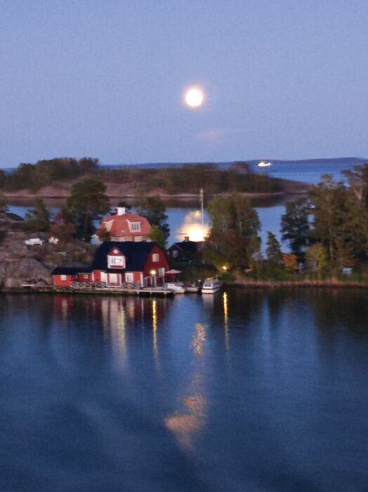 En månbelyst kustvy med träd och hus intill vattnet, fotad från Gotlandsbåten i Nynäshamn. Månen reflekteras på vattnet.