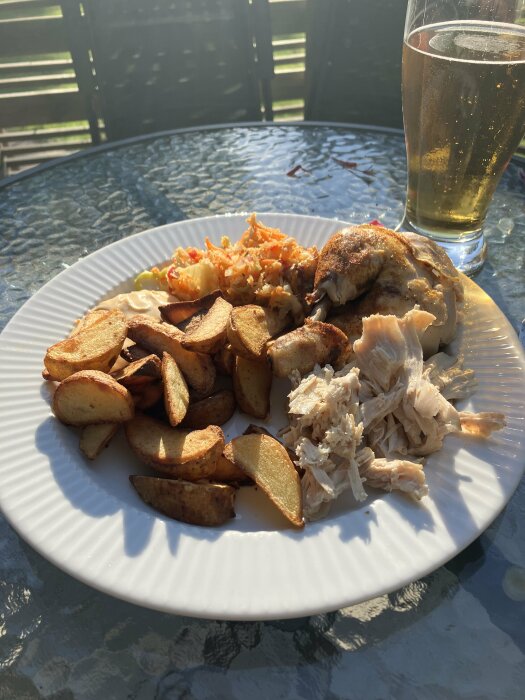 En tallrik med helstekt kyckling, klyftpotatis, råkostsallad och en sås på ett glasbord utomhus, med ett glas dryck i bakgrunden.