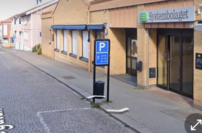 Entrén till Systembolaget med parkeringsskylt och trottoar i förgrunden, samt byggnader i bakgrunden på en landsortsgata.