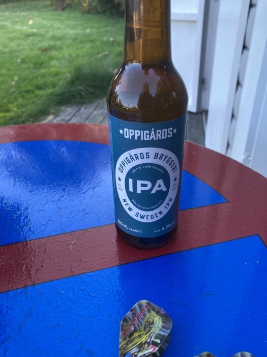 Ölflaska av märket Oppigårds IPA står på ett bord utomhus, med grön gräsmatta och byggnadsvägg i bakgrunden. Darts poängpluppar ligger på bordet.