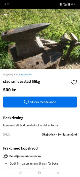 Gammalt smidesstäd på en trästubbe med omgivande gräs i bakgrunden. En järnhammare syns bredvid städet. Text: "städs smidesstäd 55kg, 500 kr.