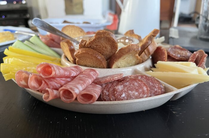 En tallrik med tapas, inklusive skruvade skivor av salami, andra charkuterier, brödkrokar, ostar och skivor grönsaker som paprika och gurka på ett bord.