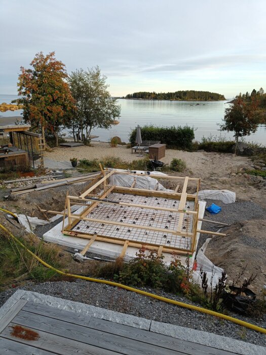 Påbörjat gjutningsarbete för nytt bad, inklädd armering och trästöd i naturskön miljö nära sjö med öar i bakgrunden och träd runtom.