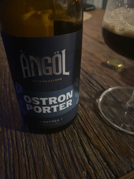 Flaska med Ängöl Ostron Porter på ett träbord bredvid ett halvtomt glas med mörk öl.