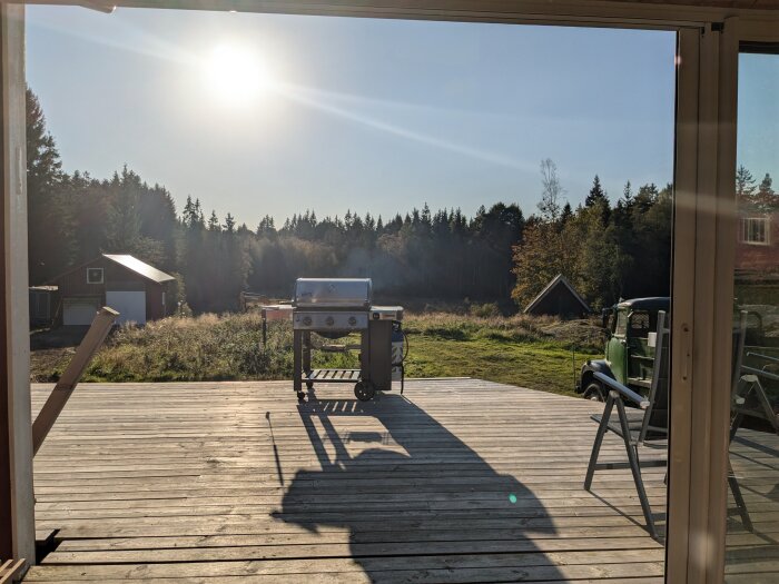 En gasgrill på en soldränkt altan med en skog och byggnader i bakgrunden under en klar sommardag.