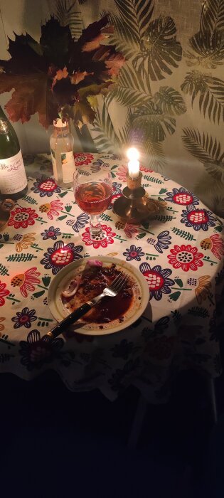 Tallrik med rester av lasagne, rödlök och vinäger bredvid en tänd ljusstake, ett vinglas och en flaska på en blommig duk.