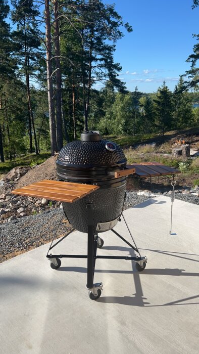 Svart keramisk grill med sidobord i trä på uteplats, omgiven av skog.