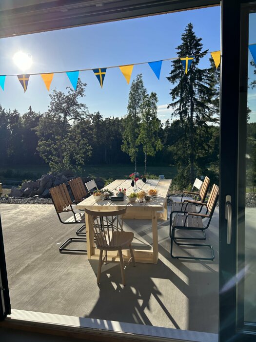 Utomhus uteplats med ett stort, tungt träbord omgivet av stolar. Bordet är dukat med mat och dryck inför midsommarfirande under blå och gul vimpeldekoration.