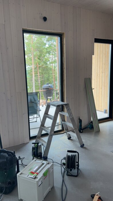 Renovering av hus med stege, sänksåg, förlängningssladd och annan utrustning på golvet. Utomhus syns en altan med grill och en stol.