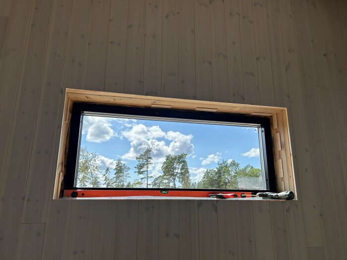 Fönster med träram och några verktyg på fönsterbrädan; utsikt med blå himmel och träd utanför.