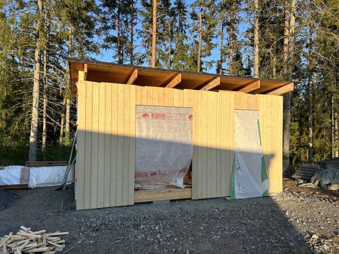 Nybyggt förråd med träpanel och råspontluckor. En del av väggen är täckt med byggplast. Skog i bakgrunden och byggmaterial i förgrunden.