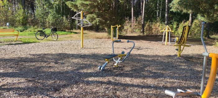 Utomhusgym med träningsredskap i en skog, en parkbänk och en cykel i bakgrunden.