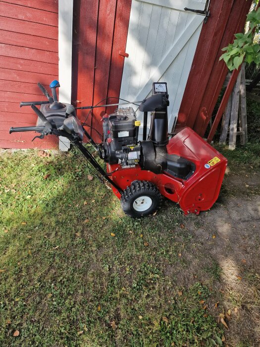 Röd snöslunga ståendes vid en röd och vit trävägg på gräsig mark, redo för vinterförberedelser.