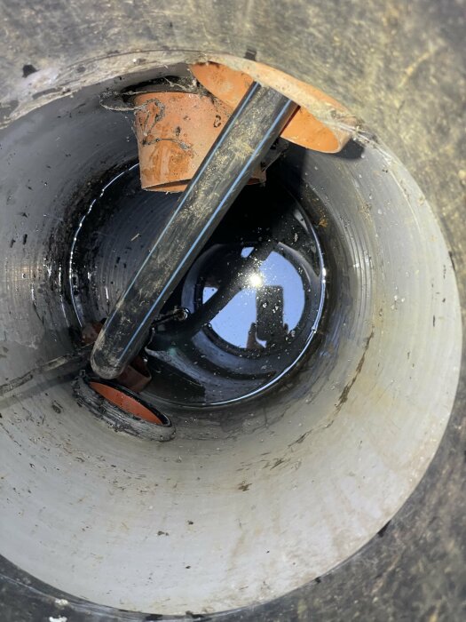 Svart rör i betongschakt, hindrar dräneringspump från att tas upp. Pumpen är under vattenytan. Orange plaströr rinner ut i schaktet.