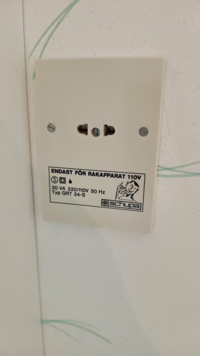 Vitt uttag för rakapparat, märkt "ENDAST FÖR RAKAPPARAT 110V", 220/110V 50Hz från Schupa. Uttaget sitter på en vit vägg med grönt mönster.