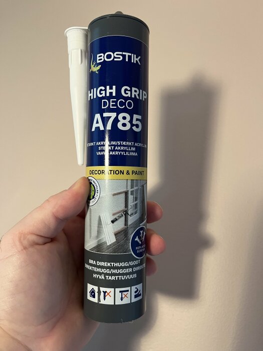 Hand som håller en tub av Bostik High Grip Deco A785 starkt akryllim med vit topp, mot en beige vägg.