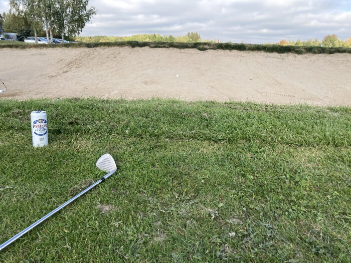 Ölburk av märket Peroni och en golfklubba placerade på gräset framför en sandbunker på en golfbana.