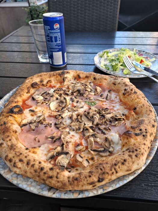 En napolitansk pizza toppad med skinka och svamp på ett bord med en tallrik sallad och en burk Pepsi i bakgrunden.