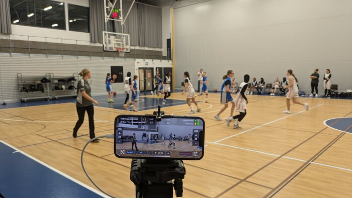 Ungdomsbasketmatch i en sporthall med spelare i blå och vita tröjor, medan en smartphone på stativ sänder matchen.