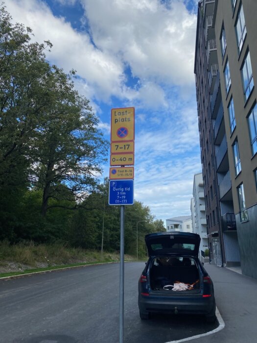 Skylt med parkeringsanvisningar för lastplats och övrig tid, parkerad bil med öppen baklucka bredvid trottoaren, höghus och träd i bakgrunden.