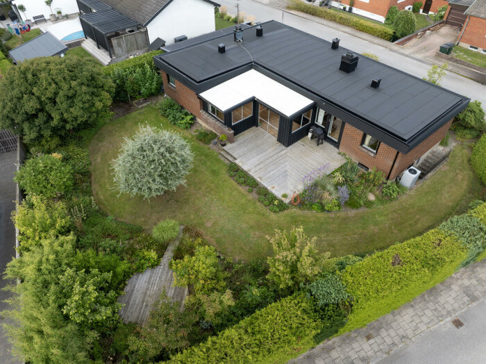 En luftig drönarbild av ett enplanshus med platt tak, byggt 1968, med trädgård och trädäck i ett bostadsområde.