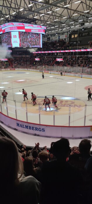 Ishockeymatch med publik i en inomhusarena, spelare i röda tröjor på isen och stor bildskärm som visar målscorer. Text och logotyper på sarg och bildskärm.