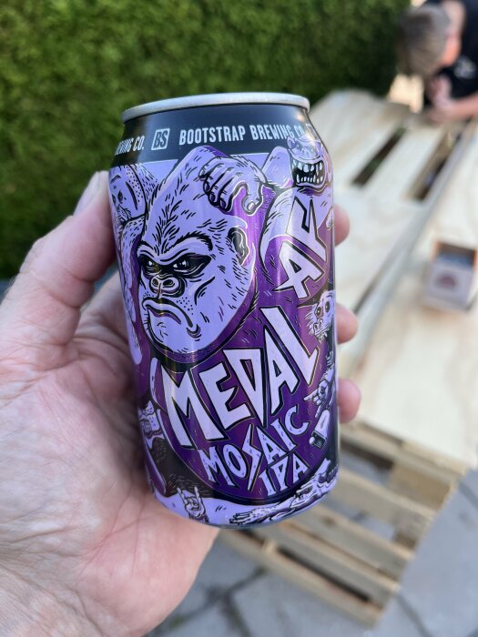 Person som håller en ölburk med texten "Medal Mosaic IPA" från Bootstrap Brewing Co. i sin hand, med en bakgrund av en trädgård och träpallar.