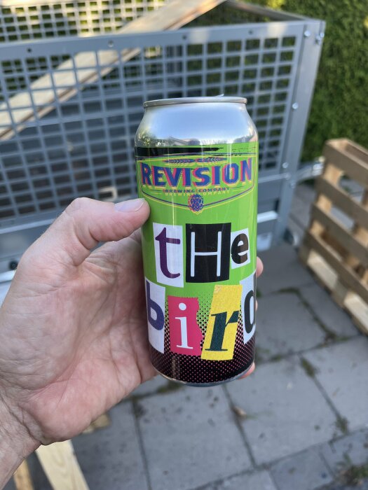 En hand håller en ölburk från Revision Brewing Company med färgglad etikett, delvis i trädgårdsmiljö och med staket i bakgrunden.