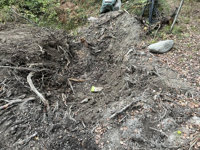 Bild av uppgrävt område i skogsmiljö, med rötter och stenar synliga i jorden. Ett stort stenblock syns till höger på marken.