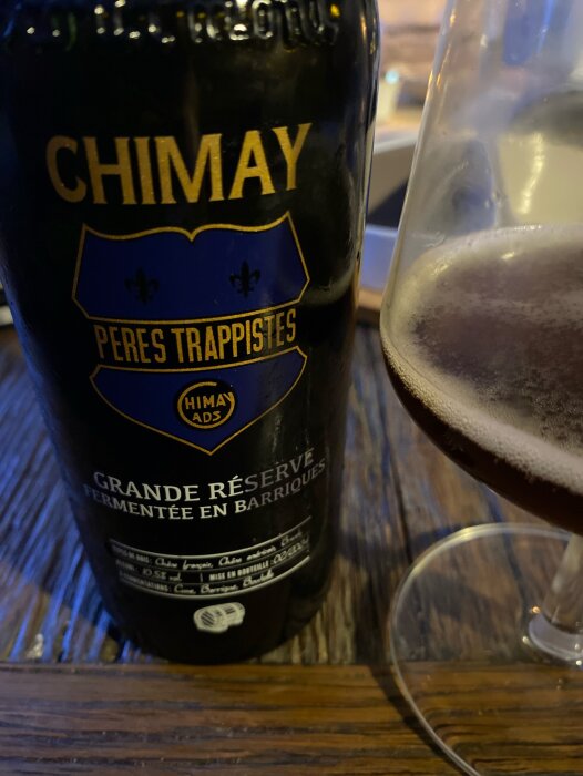 En flaska Chimay Grande Réserve öl och ett halvfullt ölglas på ett träbord.