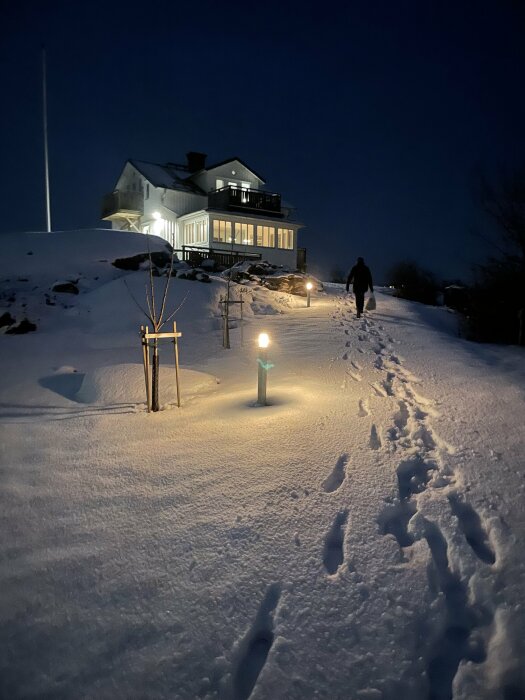 Ett upplyst hus i snölandskap på natten med flera utomhuslampor längs vägen och en person som går mot huset.