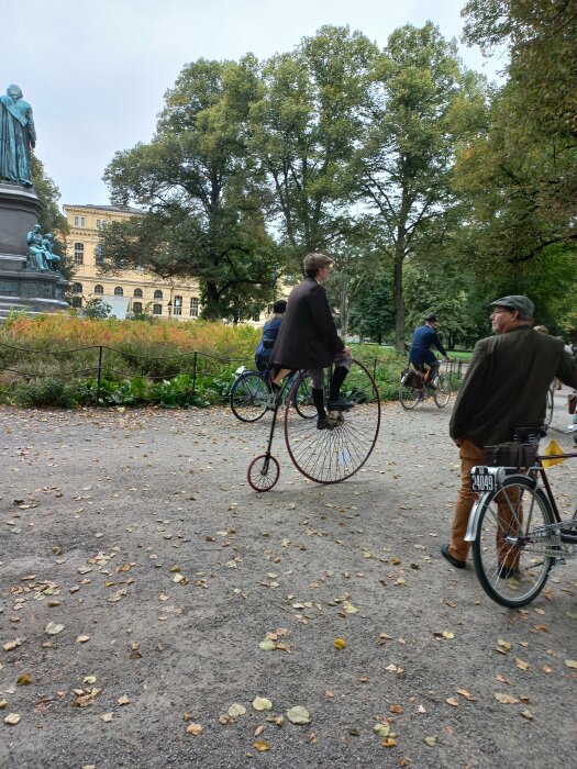 Person som cyklar på en höghjuling i en park med staty och byggnader i bakgrunden, omgiven av andra cyklister.