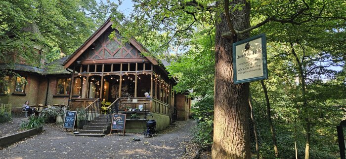 En tegelbyggnad i trä med en veranda och ljusslingor omgiven av träd. En skylt på ett träd visar texten "Queenswood Café".