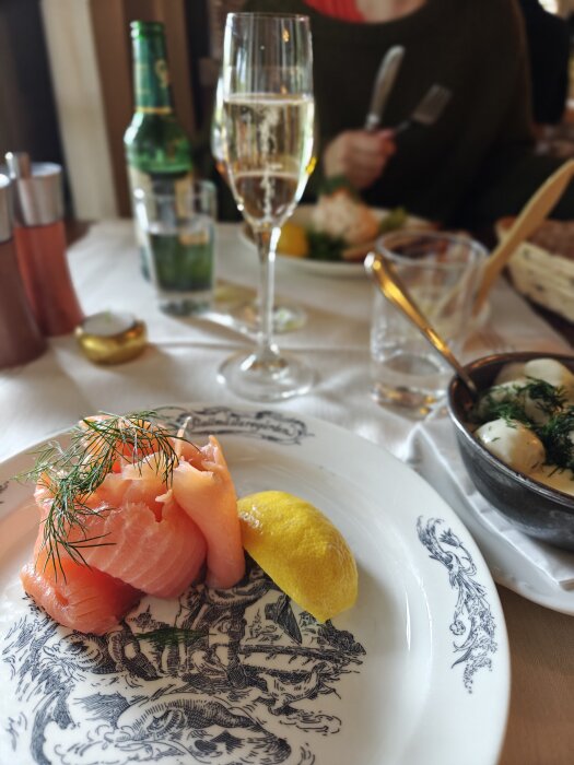 Tallrik med rimmad lax, citronskiva och dill på ett matbord med champagneglas, ölflaska och skål med potatis i bakgrunden.