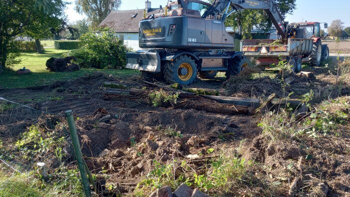 Grävmaskin och traktor arbetar med att schakta mark på en gård under utökning. Fiberduk har lagts ut och jorden förbereds för förstärkning av gårdsplanen.