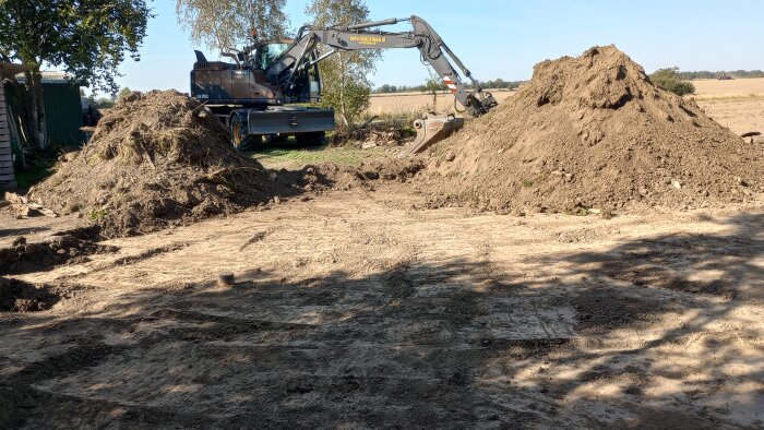 Grävmaskin i arbete med att schakta och förbereda marken med fiberduk, stora högar av jord synliga på en gårdsplan.