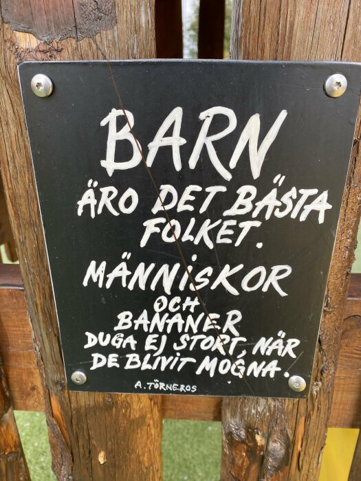 Skylt med texten "Barn äro det bästa folket. Människor och bananer duga ej stort, när de blivit mogna. A. Törneros" monterad på trä.