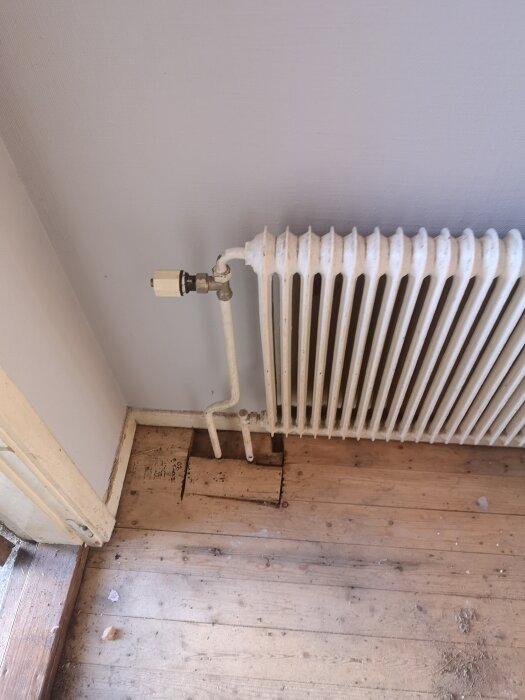 En gammal vit radiator med anslutningar syns mot en ljus vägg, placerad ovanför ett slitet trägolv.